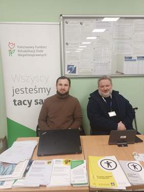 Pokaż zdjęcie: Dwóch mężczyzn siedzi przy stole z ulotkami oraz broszurami. Za nimi tablica informacyjna oraz baner z logo PFRON