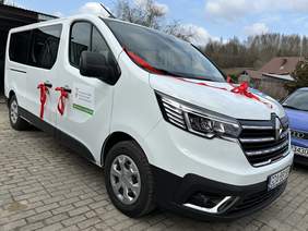 Pokaż zdjęcie: Biały mikrobus marki Renault. Z czerwonymi kokardami przy klamkach i na środku maski. Na drzwiach naklejka z informacją o dofinansowani ze środków PFRON i logo Funduszu.