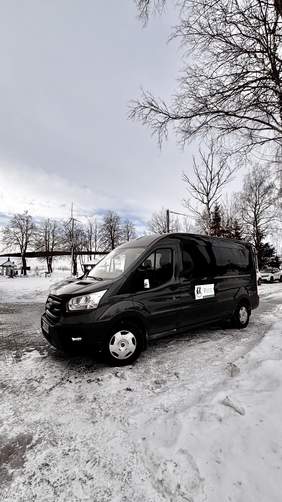 Pokaż zdjęcie: Ciemnoszary dziewięciomiejscowy mikrobus przystosowany do przewozu osób z niepełnosprawnościami zaparkowany przed budynkiem w zimowej scenerii. Na drzwiach pojazdu widoczna tablica informująca o dofinansowaniu ze środków PFRON.