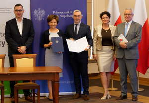 Grupa uczestników podpisania umowy stoi na tle fal Polski i roll-upów PFRON i Ministerstwa Rodziny, Pracy i Polityki Społecznej.