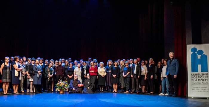 Pokaż zdjęcie: Na cenie w dwóch rzędach stoi bardzo duża grupa osób pozując do pamiątkowego zdjęcia. Na podłodze po turecku, przed tymi osobami siedzi ksiądz oraz siostra zakonna. Obok księdza stoi duży kosz kwiatów, z boku zdjęcia baner logo hospicjum małego Księcia w Lublinie oraz informacja z prośbą o przekazanie 1% podatkuz informacją o 