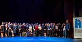 Pokaż zdjęcie: Na cenie w dwóch rzędach stoi bardzo duża grupa osób pozując do pamiątkowego zdjęcia. Na podłodze po turecku, przed tymi osobami siedzi ksiądz oraz siostra zakonna. Obok księdza stoi duży kosz kwiatów, z boku zdjęcia baner logo hospicjum małego Księcia w Lublinie oraz informacja z prośbą o przekazanie 1% podatkuz informacją o 