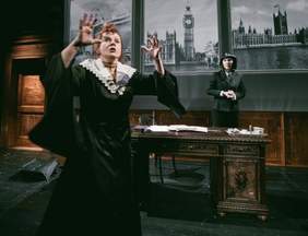 Pokaż zdjęcie: Fragment sceny na której grane jest przedstawienie. Jedna aktorka stoi za drewnianym biurkiem i przygląda się kobiecie stojącej przed nią. 