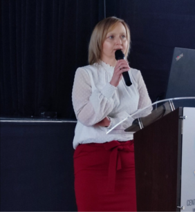 Pokaż zdjęcie: Przy mównicy kobieta w białej bluzce i bordowej spódnicy.