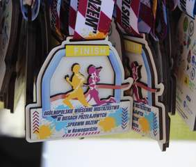 Pokaż zdjęcie: Zbliżenie na zawieszone na sznurkach medale z kolorowymi wstążkami. Medale mają kształt łuku z sylwetkami biegnących osób i napisem „FINISH” na górze. Poniżej znajduje się tekst: „Ogólnopolskie Wiosenne Mistrzostwa w Biegach Przełajowych – Sprawni Razem – w Nowogardzie”. Tło medali jest ozdobione dynamicznymi kolorami i grafikami sportowymi.