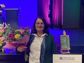 Pokaż zdjęcie: Kobieta w eleganckiej marynarce stoi na scenie z bukietem kwiatów i statuetką ustawioną na postumencie obok. Za nią kolorowe światła oraz dekoracje gali.