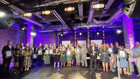 Pokaż zdjęcie: Na scenie stoi kilkadziesiąt osób: kobiet i mężczyzn. Tworzą dwa rzędy. Trzymają w rękach dyplomy, statuetki, kwiaty. Wszyscy są elegancko ubrani. 