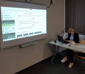Pokaż zdjęcie: Sala szkoleniowa z dużym ekranem projektora po lewej stronie, na którym wyświetla się widok aplikacji iPFRON+. Po prawej stronie, przy białym stole, siedzi pracownik Oddziału Wielkopolskiego PFRON z otwartym laptopem.