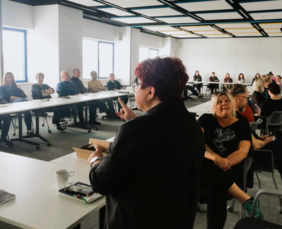 Uczestnicy spotkania siedzą przy stołach, a na pierwszym planie tłumaczka języka migowego