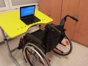 Biurko, na którym stoi laptop, dostosowane dla osób poruszających się na wózku inwalidzkim, pochylone, z zabezpieczeniem przed spadaniem przedmiotów, dostosowane do podjazdu wózkiem