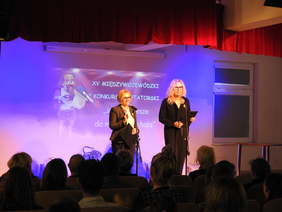 Pokaż zdjęcie: Dwie kobiety ubrane w ciemnych eleganckich strojach. Obie w blond włosach jedna w krótszych. Obie mają na sobie okulary. Stoją przed mikrofonami mówią uśmiechając się.