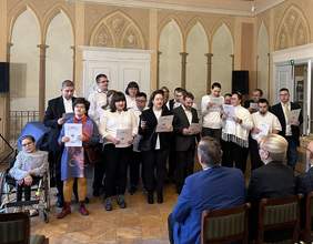 Pokaż zdjęcie: W sali muzeum na ścianach gdzie namalowane są zabytkowe łuki na środku stoi grupa ludzi elegancko ubranych w białe koszule i marynarki. Wszyscy trzymają tekst w rękach i śpiewają. Naprzeciw na krzesłach siedzą goście.
