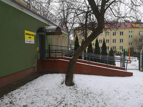 Pokaż zdjęcie: Długi podjazd dla wózków prowadzący do drzwi wejściowych do ośrodka, z boku na zielonej elewacji żółta tabliczka informacyjna z treścią, Ośrodek Wsparcia i Testów w Świdniku, poniżej mało logo OWIT oraz PFRON