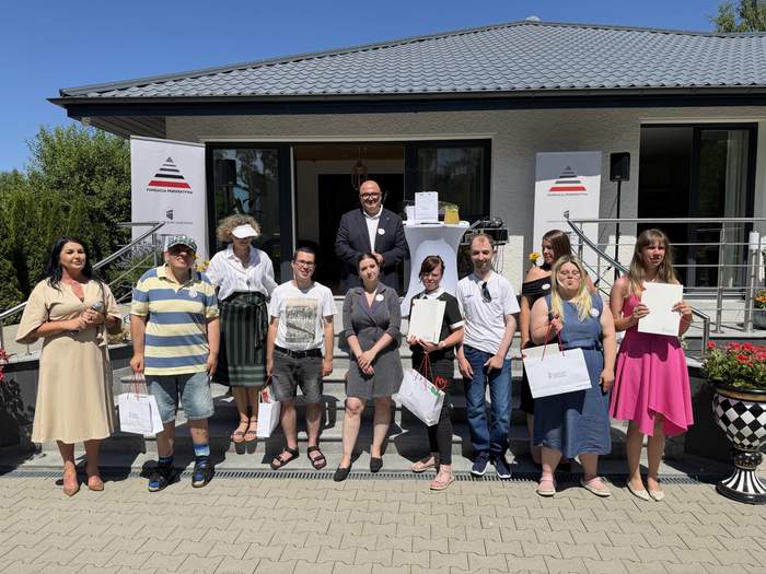 Pokaż zdjęcie: Grupa osób pozuje przed wejściem do budynku. Część osób trzyma dyplomy i torby z upominkami. W tle widoczne są schody z poręczami oraz wejście do nowoczesnego domu parterowego.