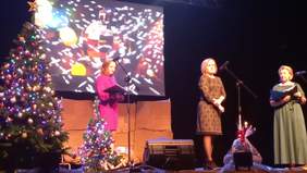 Pokaż zdjęcie: Na scenie przy mikrofonie stoi starsza kobieta ubrana w czarną sukienkę we wzory. Obok niej stoi kobieta w długiej zielonej sukni. Na scenie są świąteczne dekoracje, dwie choinki. W tle ekran z prezentacją  