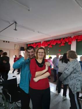 Pokaż zdjęcie: Na zdjęciu zbliżenie na parę tańczącą podczas Balu Walentynkowego, para trzyma się za ręce, spogląda w kierunku obiektywu. W tle widać inne tańczące osoby.