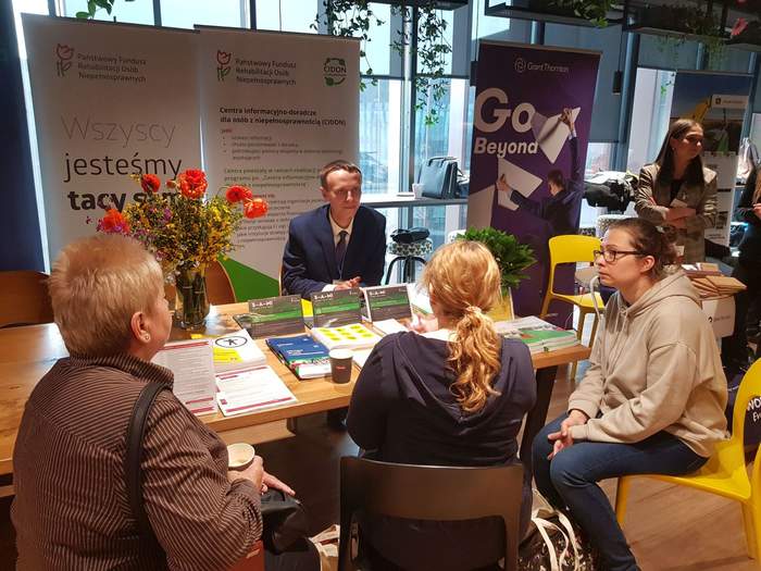 Pokaż zdjęcie: Na Targach Pracy, przy stoisku Oddziału Wielkopolskiego PFRON siedzą trzy kobiety, którym mężczyzna w garniturze udziela informacji. W tle widoczne są dwa banery: CIDON oraz PFRON. Na stoliku ułożone zostały trzy bukiety kwiatów oraz materiały informacyjne promujące programy i zadania PFRON. W tle widoczne są także stoiska innych pracodawców.