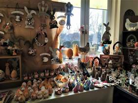 Pokaż zdjęcie: Ozdoby wielkanocne, w tym między innymi: zające, kury, baranki, czy też aniołki ułożone zostały na blacie stołu, parapecie okiennym oraz na kilku półkach. Część ozdób jest podwieszona na drewnianej belce przymocowanej do ściany.