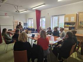 Pokaż zdjęcie: Kilkanaście osób siedzi przy dużym stole. Przed nimi mężczyzna, który omawia programy realizowane przez PFRON. Znajdują się w dużej sali.