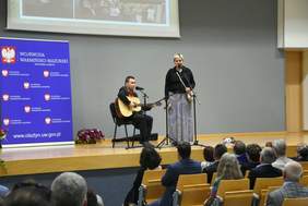 Pokaż zdjęcie: Na scenie gitarzysta i wokalistka z tamburynem śpiewają do mikrofonu