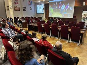 Duża sala, widok na część zebranych z tyłu, bokiem oraz przed nimi widać grupę siedzących dyskutujących osób, nad którymi jest ekran. Na ekranie wyświetlają się osoby biorące udział w dyskusji.