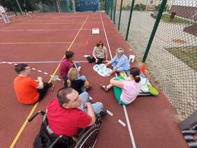 Pokaż zdjęcie: Grupa osób, w tym osoba na wózku, siedzi na boisku sportowym w kręgu i bierze udział w zajęciach z elementami gier i ćwiczeń integracyjnych.