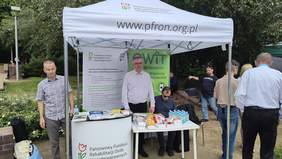 Pokaż zdjęcie: Namiot 2x2 w kolorze białym z logiem PFRON. W tle widoczne dwa banery PFRON i OWiT. Widoczni dwaj pracownicy pozujący do zdjęcia. 