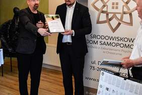 Pokaż zdjęcie: Grupa mężczyzn stoi we wnętrzu sali, gdzie odbywa się oficjalne wydarzenie. Dwóch z nich trzyma wspólnie dyplom, który jeden z nich prezentuje. W tle znajdują się banery z logo i nazwą „Środowiskowy Dom Samopomocy dla Osób z Zaburzeniami Psychicznymi w Choszcznie”. Mężczyźni są elegancko ubrani, trzymają także torby prezentowe i plansze edukacyjne. Wnętrze jest ciepłe, drewniana podłoga i dekoracyjne elementy tworzą przytulną atmosferę.