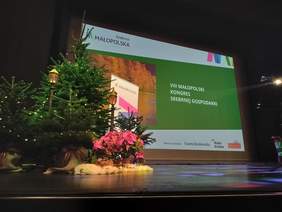 Pokaż zdjęcie: Scena w Operze Krakowskiej. Na scenie dekoracje świąteczny, choinki oraz ekran. Na zielonym tle biały napis: VIII Małopolski Kongres Srebrnej Gospodarki