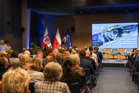 Pokaż zdjęcie: Szerokie ujęcie sali konferencyjnej, na którym widać zgromadzoną publiczność, siedzącą rzędami tyłem do obiektywu. Na podwyższeniu, w głębi sali, stoi mężczyzna w garniturze za mównicą, przemawiający do zebranych. Za mównicą umieszczone są dwie flagi Polski i flaga Unii Europejskiej. Po prawej stronie, na dużym ekranie, wyświetlana jest prezentacja z hasłem: "NOWOCZESNE TECHNOLOGIE JAKO ŹRÓDŁO ZMIAN W EDUKACJI ZAWODOWEJ". W prawym górnym rogu ekranu widać logo "Pomorze Zachodnie" i inne logotypy.