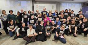 Pokaż zdjęcie: Na środku sali grupa osób - laureatów konkursu plastycznego. Wszyscy ubrani są w czarne koszulki na których jest nadruk nagrodzonych prac plastycznych.