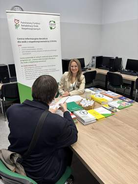 Pokaż zdjęcie: Konsultacja przy stoisku informacyjnym. Kobieta w jasnym żakiecie rozmawia z mężczyzną siedzącym tyłem. Na stole leżą materiały informacyjne i koszyk z cukierkami. W tle stoi roll-up z napisem: „PFRON – Centra informacyjno-doradcze dla osób z niepełnosprawnością”.