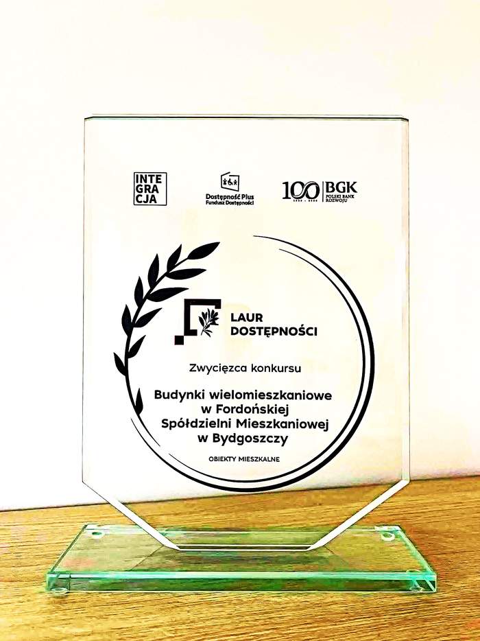 Szklana statuetka w kształcie proporczyka na podtsawie. Na niej widnieja napisy: u góry od lewej -  logo Integracja, logo Funduszu Dostepności Dostępność plus, logo Polskiego Banku Rozwoju BGK. Poniżej, centralnie: w kole, które tworzy gałązka liścia laurowego: logo konkursu, pod spodem napis Zwycięzca konkursu Budynki wielomieskzaniowe w Fordońskiej Spółdzielni Mieszkaniowej w Bydgoszczy Budynki mieszkalne