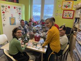 Pokaż zdjęcie: Grupa osób siedzi przy stole, dekorując stroiki świąteczne. Kobieta w okularach trzyma doniczkę z gałązkami, obok niej stoi uśmiechnięta dziewczyna w pomarańczowym swetrze. W tle pozostali uczestnicy skupieni na pracy. Pomieszczenie jest kolorowe, z ozdobami na ścianach i oknach.