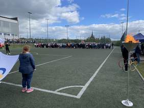 Pokaż zdjęcie: Na sztucznej murawie dużego boiska piłkarskiego stoi wielu zawodników. Po lewej stronie kobieta trzymająca w ręku flagę z logo igrzysk olimpijskich po prawej płonący znicz olimpijski.