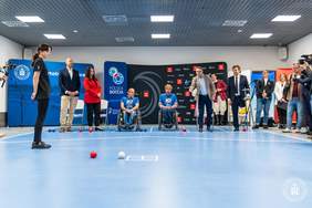 Pokaż zdjęcie: Grupa kilkunastu osób znajduje się na boisku do gry w Boccie, a wśród tych osób jest jeden mężczyzna, który wrzuca na boisko jedną bilę w kolorze niebieskim. W tle ustawione są dwie ścianki promujące Politechnikę Poznańską oraz Polską Boccie.