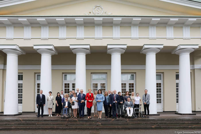 Pokaż zdjęcie: Grupa osób pozuje do zdjęcia, za nimi znajduje się biało-kremowy budynek z sześcioma kolumnami
