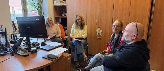 Pokaż zdjęcie: Cztery osoby siedzą w biurze przy biurku z komputerem i dokumentami; za nimi drewniane szafy i okno z widokiem na budynek oraz drzewo. Atmosfera rozmowy wygląda na spokojną i roboczą.