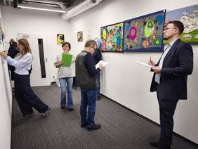 Cztery osoby oceniają prace artystyczne wiszące na ścianie w korytarzu. Dwie kobiety i dwóch mężczyzn trzymają notatniki i długopisy, studiując kolorowe obrazy i tkaniny, w tym jedną w żywych, abstrakcyjnych kolorach i inną przedstawiającą motywy samochodów i balonów.