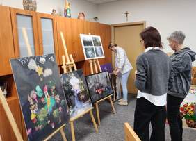 Pokaż zdjęcie: Trzy kobiety oglądają prace plastyczne ustawione na sztalugach. Na obrazach widać kolorowe, fantazyjne kompozycje oraz fotografie. Jedna z kobiet poprawia jedną z prac, a pozostałe dwie przyglądają się uważnie ekspozycji.
