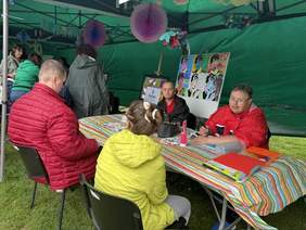 Pokaż zdjęcie: Osoby siedzą przy stole pod zielonym namiotem, uczestnicząc w warsztatach artystycznych. Na stole leżą przybory plastyczne, a w tle widać kolorowe prace w stylu pop-art.