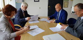 Cztery osoby siedzące przy jasnym stole, podpisujące umowę, z lewej kobieta i mężczyzna, z prawej dwóch mężczyzn