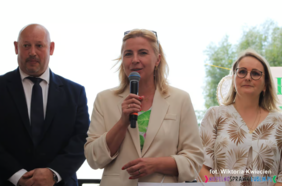 Pokaż zdjęcie: Zdjęcie trzech osób, mężczyzna w garniturze oraz dwie kobiety. Kobieta w jasnym garniturze trzyma w ręku mikrofon.