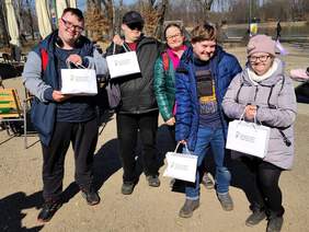 Pokaż zdjęcie: Uśmiechnięte kobiety i mężczyźni z zespołem Downa. W rękach trzymają torebki z logo PFRON