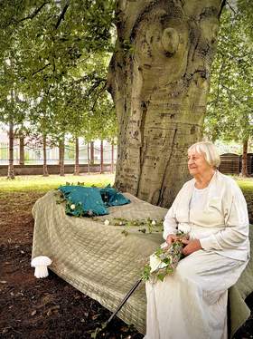 Fotografia przedstawiająca kobietę w bieli siedzącą na ławce w parku i patrzącą w dal
