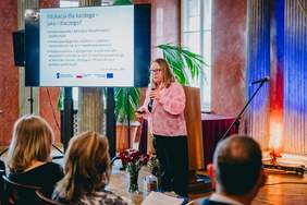 Pokaż zdjęcie: Sala konferencyjna z publicznością siedzącą na krzesłach i zwróconą w stronę sceny. Na środku stoi osoba przemawiająca do publiczności i trzyma mikrofon. Po lewej stronie sceny ustawiony jest ekran projekcyjny i wyświetlony jest na nim slajd. Ściany mają ciepły, czerwony kolor, a oświetlenie w odcieniach pomarańczowo niebieskich podkreśla kolumny. Na ekranie projekcyjnym widnieje tekst: „Edukacja dla każdego – jaka i dlaczego”. Niżej, mniejszym drukiem wypunktowano: zmiana wyrosła z koncepcji filozoficznych i społecznych, zmiana paradygmatu myślenia o zjawisku różnorodności, zmiana myślenia o dostępności kształcenia dla wszystkich uczniów w kierunku zmiany istniejącej infrastruktury i postaw społecznych.