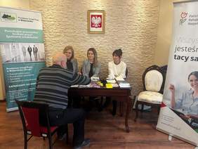 Pokaż zdjęcie: Przy drewnianym stole siedzą trzy kobiety – dwie ubrane w marynarki i jedna w jasnym swetrze. Naprzeciwko nich, na krześle, siedzi starszy mężczyzna, prowadząc rozmowę. Na stole znajdują się ulotki, długopisy i materiały informacyjne. Po lewej stronie widoczny jest zielony baner Powiatowego Urzędu Pracy w Białymstoku, a po prawej biały baner PFRON z napisem „Wszyscy jesteśmy tacy sami”. W tle, na ścianie, wisi godło Polski.