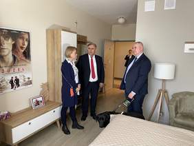 Pokaż zdjęcie: W pokoju z jasnymi ścianami i jasnymi meblami stoi i rozmawia kobieta i dwóch mężczyzn. Wszyscy ubrani na galowo. Jeden z mężczyzn trzyma na uprzęży psa przewodnika – ciemny labrador. Po lewej stronie plakat na ścianie z filmu Żużel.