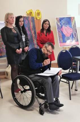 Gratulacje dla PZN-u składa Pan Krzysztof Kranz - przedstawiciel Prezydenta Bydgoszczy, siedzi na wózku inwalidzkim , na kolanach trzyma pismo, a w dłoni mikrofon, za nim stoi Pani Prezez PZN-u Okręgu Kujawsko-Pomorskiego oraz dwie kobiety