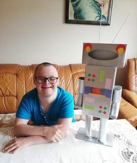 Pokaż zdjęcie: Uczestnik zajęć siedzi przy stole, pozuje z kartonowym robotem - praca wykonana w ramach konkursu "Coś niezwykłego z niczego"